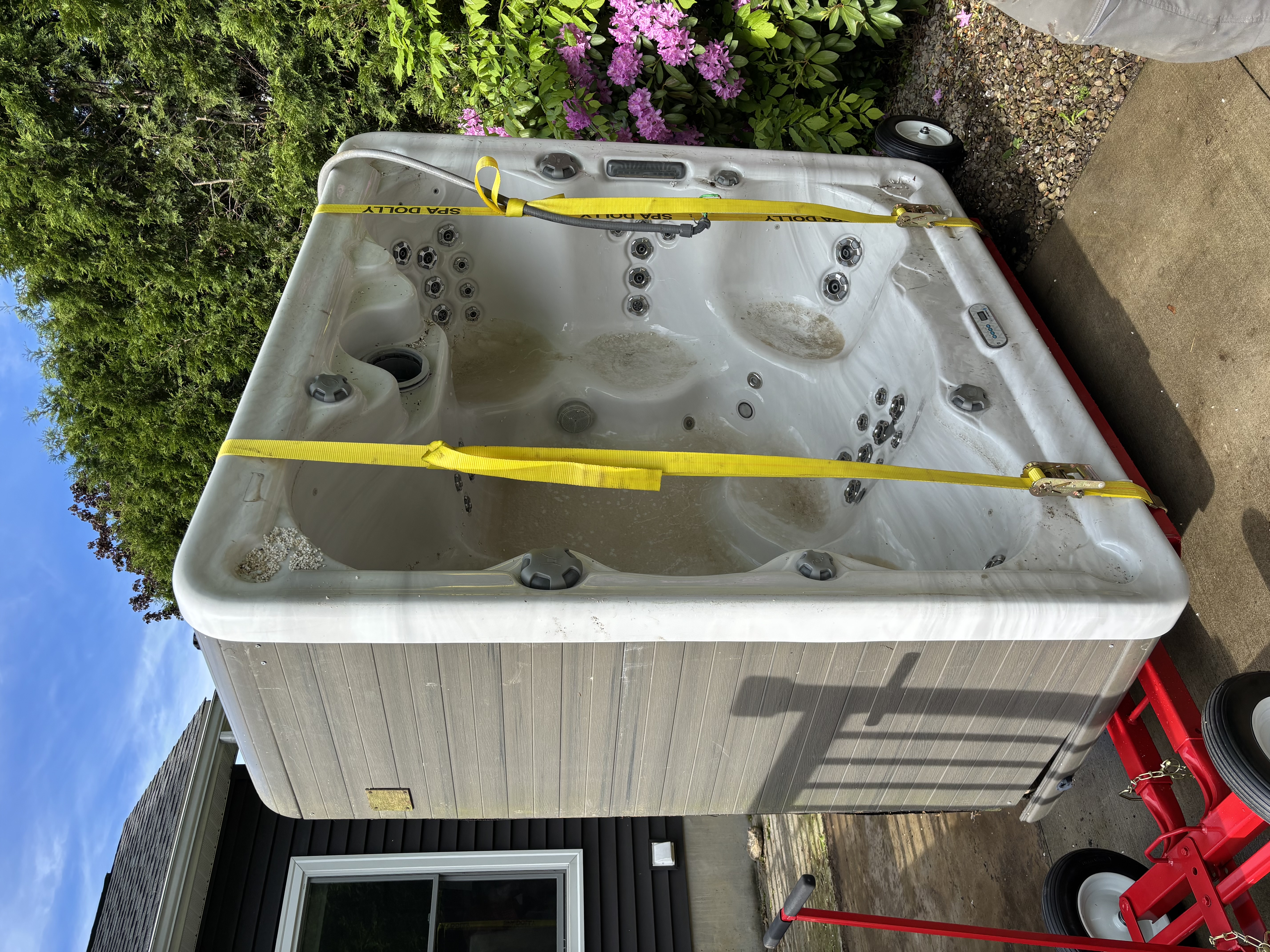 Hot tub strapped upright on a red trailer beside a house during pickup